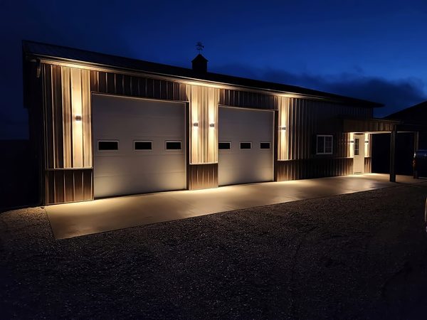 Steel shop at night with up-and-down wall sconces washing the siding between the overhead doors